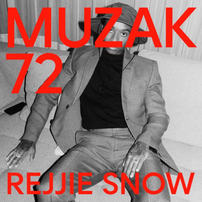 Man sitting on a sofa wearing a suit and hat. The image is black and white.