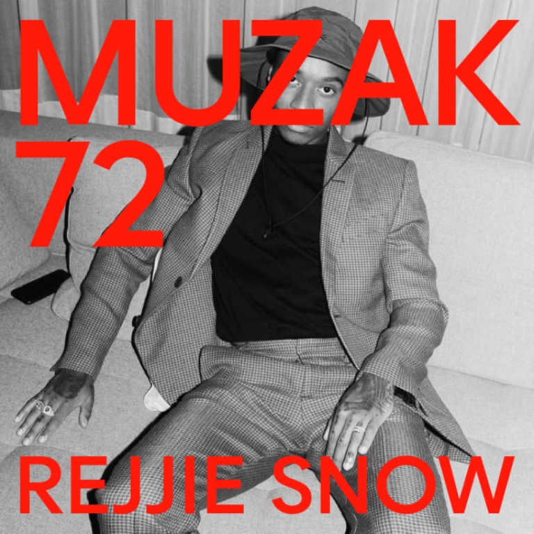 Man sitting on a sofa wearing a suit and hat. The image is black and white.